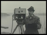 En man i rock och hatt bredvid en mätutrustning monterad på ett tripod stativ.